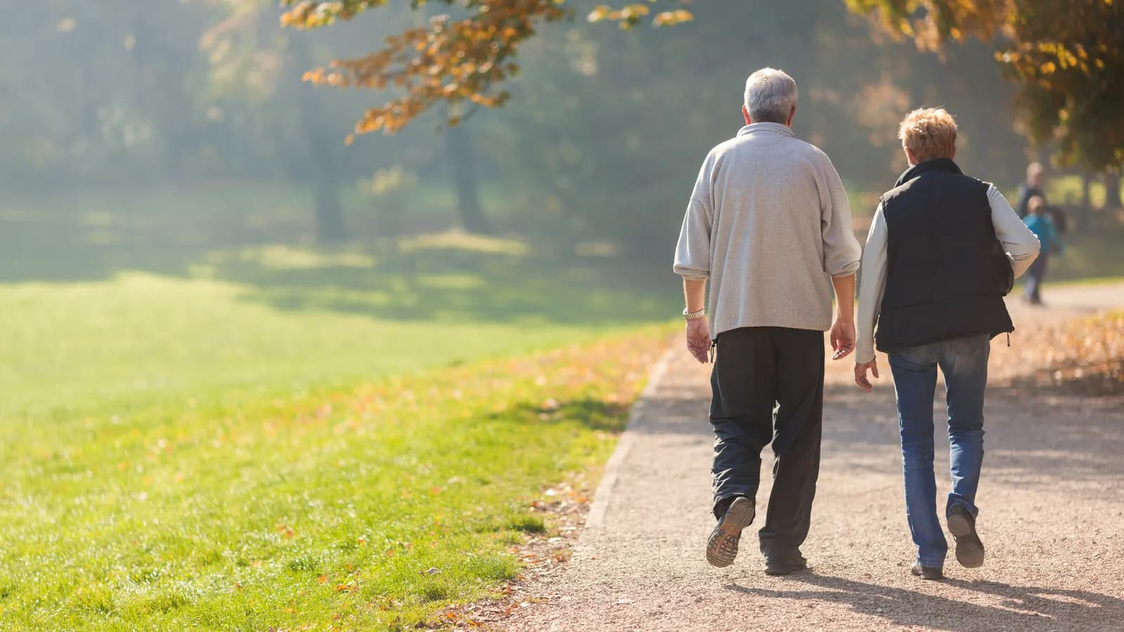 Two older adults walking along a park path, seen from behind.